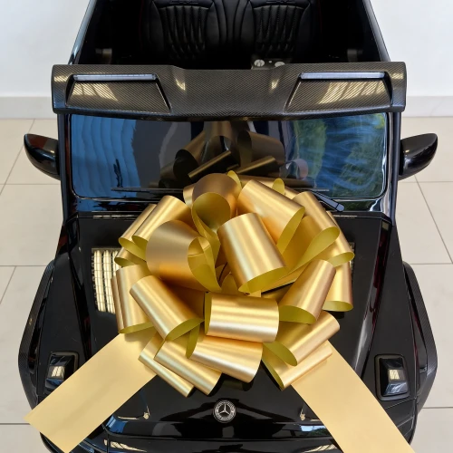 Złota kokarda prezentowa na masce, emblemat Mercedesa i karbonowa listwa dachowa z oświetleniem LED – zbliżenie czarnego Mercedesa AMG G63 XXL na akumulator dla dzieci.webp